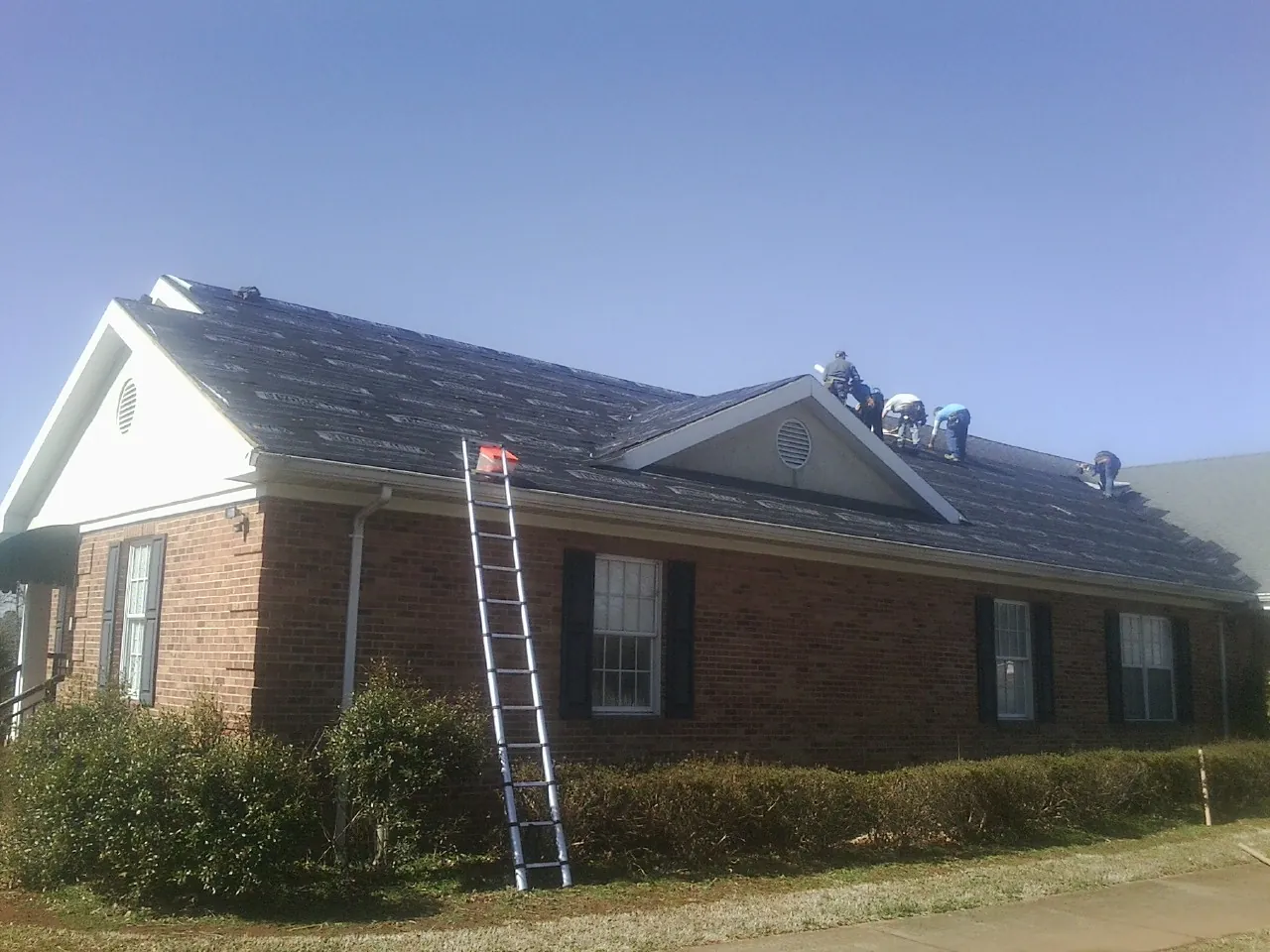 Crew tearing shingles off a brick ranch roof with a ladder set — Emergency Roof Repair work by our crew in Pratts