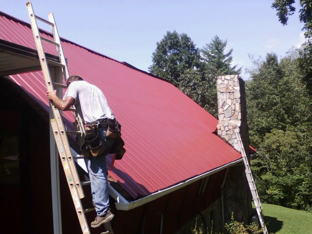 Roofer on a ladder beside a red metal roof and stone chimney — Soffit & Fascia Repair by Roofing Brothers in Pratts